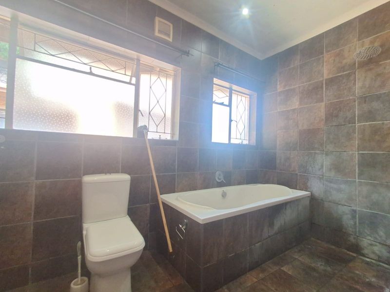 A functional bathroom featuring dark tiled walls and flooring, a built-in bathtub, and a toilet, with natural light provided by two windows.
