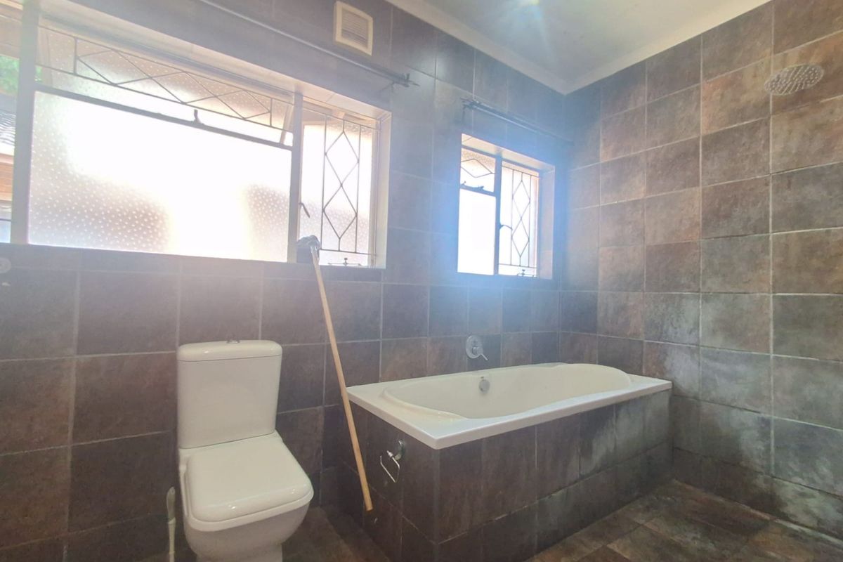 A functional bathroom featuring dark tiled walls and flooring, a built-in bathtub, and a toilet, with natural light provided by two windows.