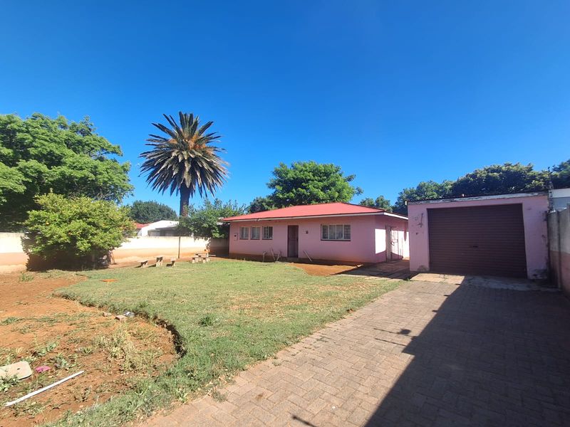 Rear view of a single-story house featuring a metal roof, attached garage with roller door, paved driveway, and a spacious garden area.