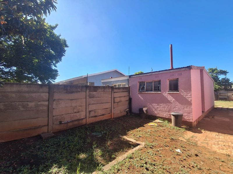 Rear exterior view of the property featuring a pink brick structure, a precast concrete boundary wall, and a small garden area requiring maintenance.
