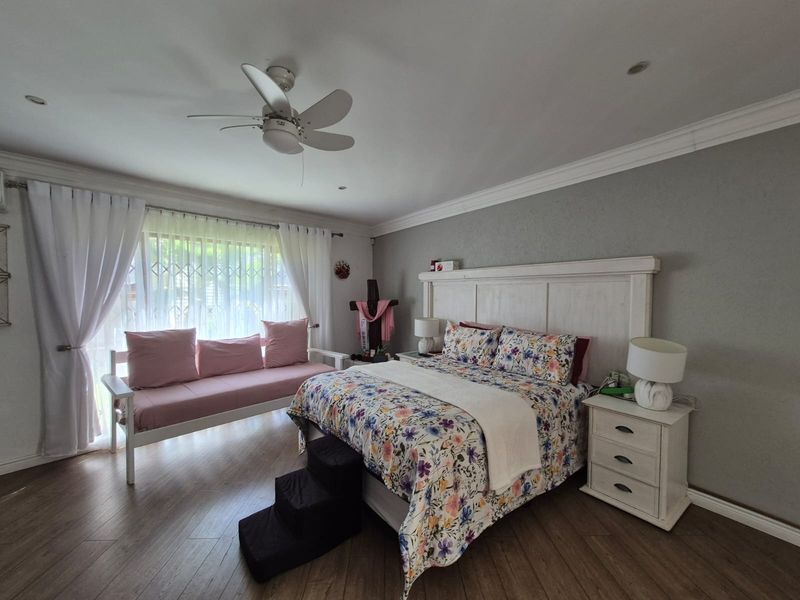 A spacious bedroom featuring laminate flooring, crown molding, recessed lighting, and a ceiling fan, with a large window protected by a security gate.