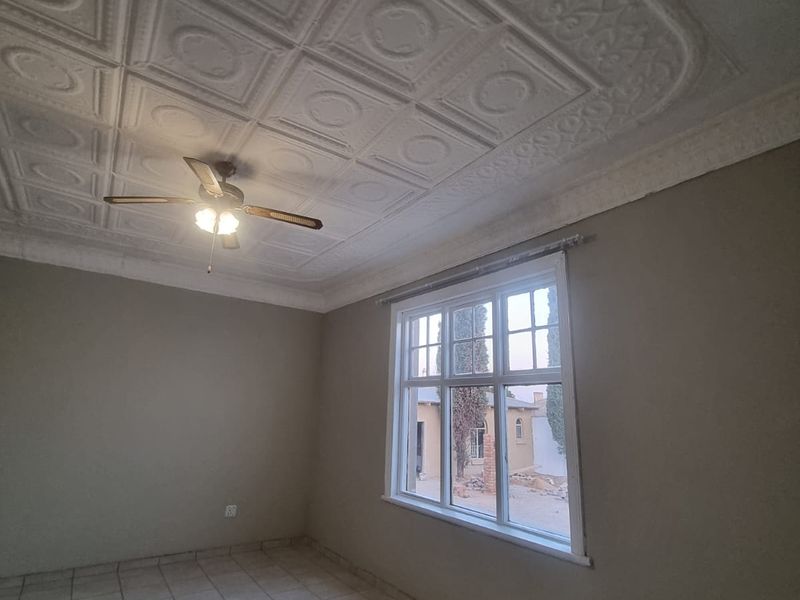 An empty bedroom featuring decorative ceiling tiles, a ceiling fan, tiled flooring, and a large window providing natural light.