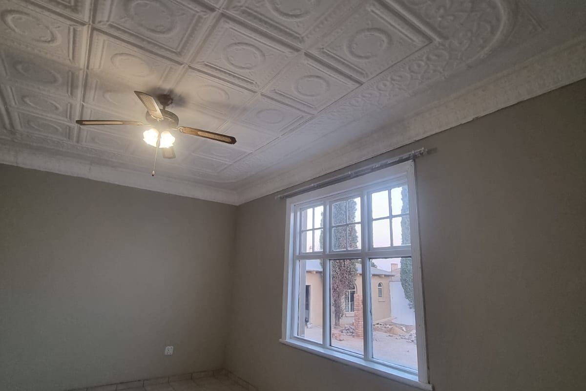 An empty bedroom featuring decorative ceiling tiles, a ceiling fan, tiled flooring, and a large window providing natural light.