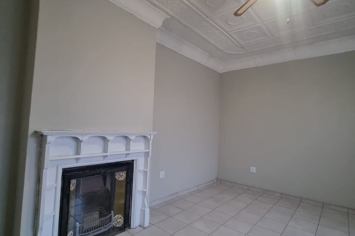 An empty living room featuring a classic fireplace with a white mantelpiece, ornate pressed metal ceilings, and neutral tiled flooring.