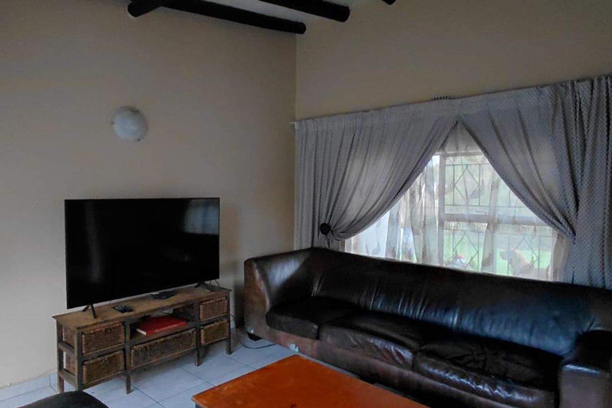 A living room featuring exposed ceiling beams, tiled flooring, and large windows fitted with security burglar bars.