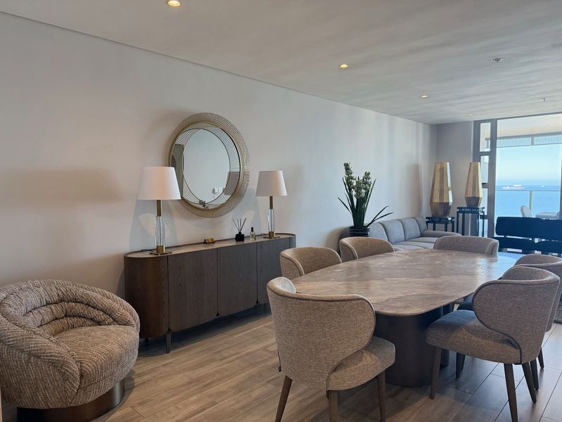 A modern open-plan living and dining area featuring neutral tones, recessed lighting, and large sliding doors leading to a balcony with ocean views.