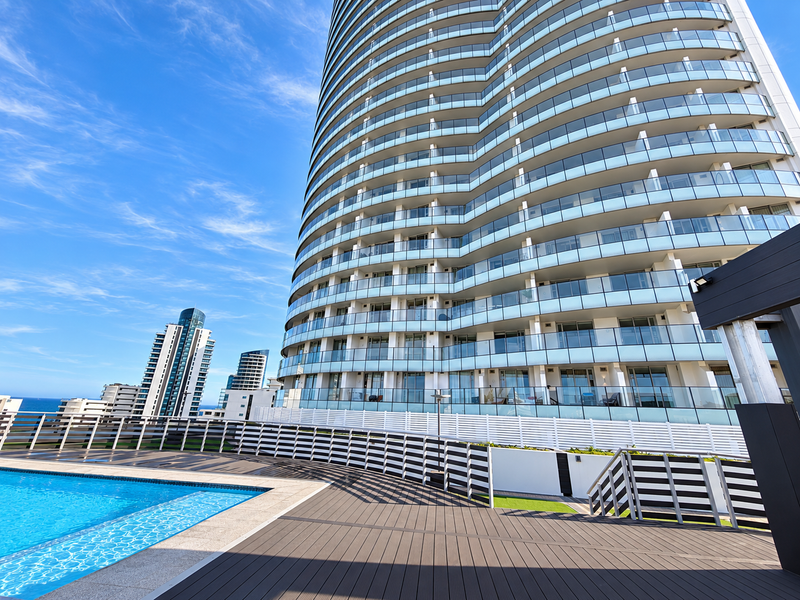 A modern high-rise apartment building exterior featuring a swimming pool, composite decking, and glass-fronted balconies with clear blue sky views.