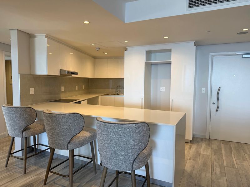 Modern open-plan kitchen featuring sleek white cabinetry, stone countertops, a breakfast bar, and integrated appliances, finished with contemporary tiled flooring and recessed lighting.