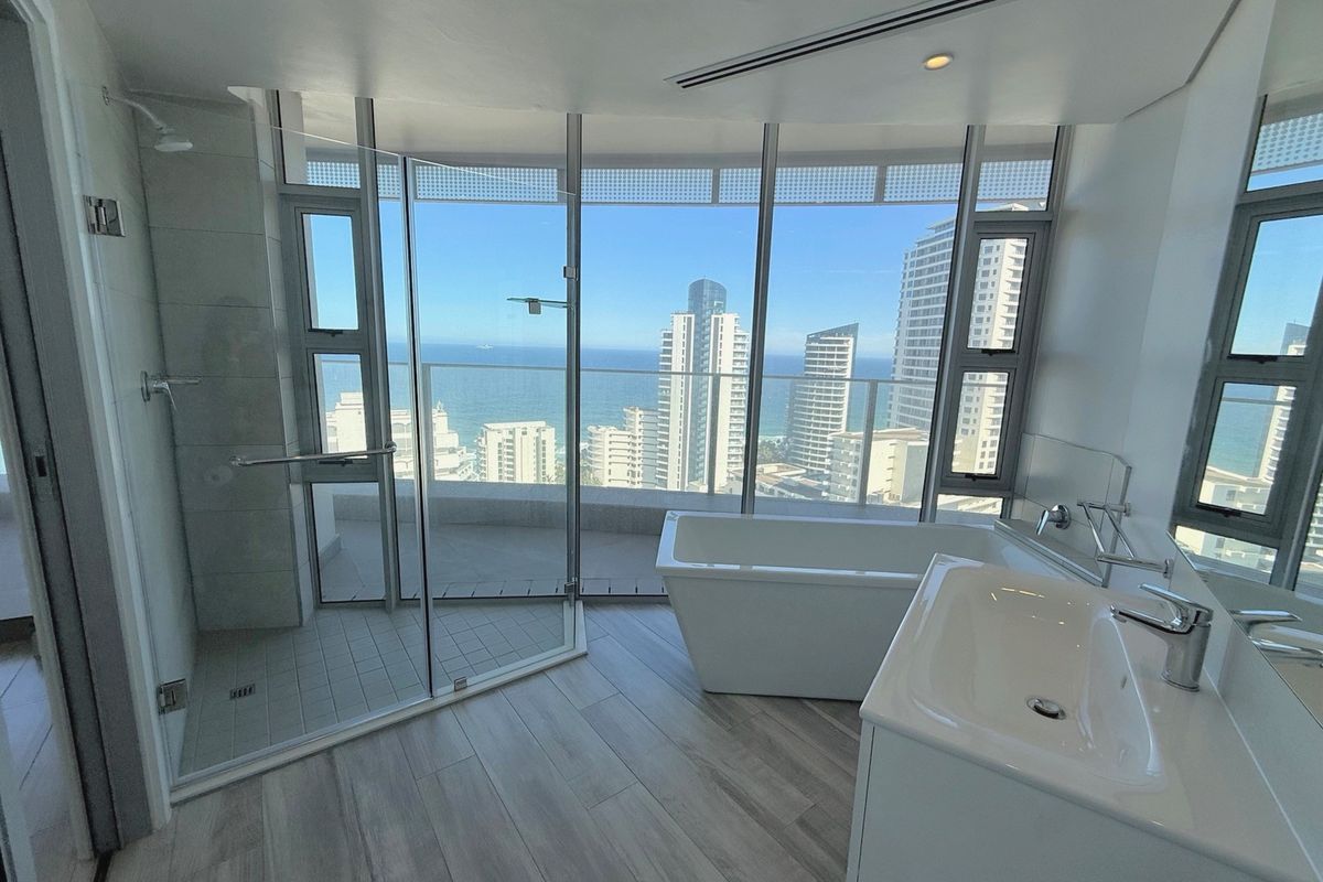 Modern bathroom featuring a freestanding bathtub, glass-enclosed shower, and vanity basin, complemented by floor-to-ceiling windows offering expansive coastal and city views.