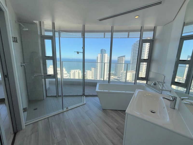 Modern bathroom featuring a freestanding bathtub, glass-enclosed shower, and vanity basin, complemented by floor-to-ceiling windows offering expansive coastal and city views.