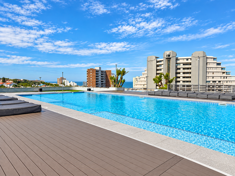 A well-maintained rooftop swimming pool area featuring modern composite decking, stone coping, and panoramic urban and coastal views.