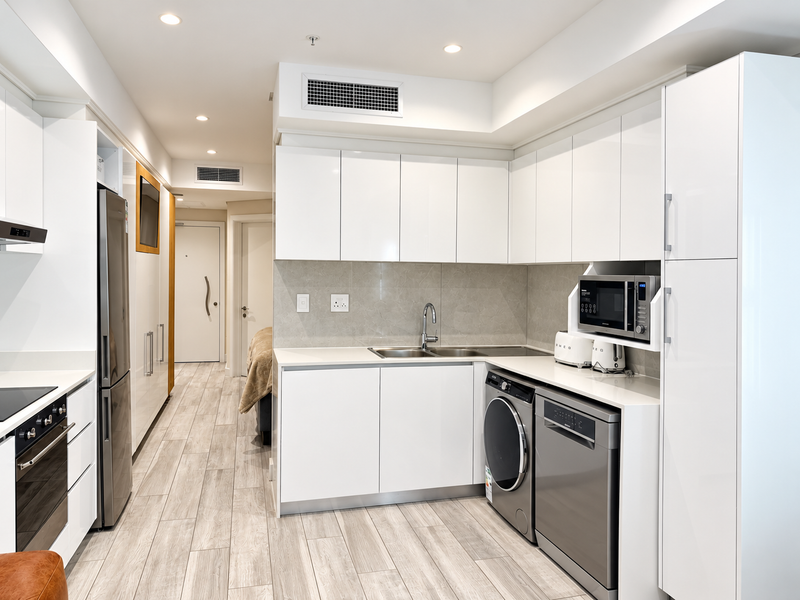 Modern kitchen featuring sleek white cabinetry, stone countertops, integrated appliances, and high-quality tiled flooring, seamlessly flowing into the apartment hallway.