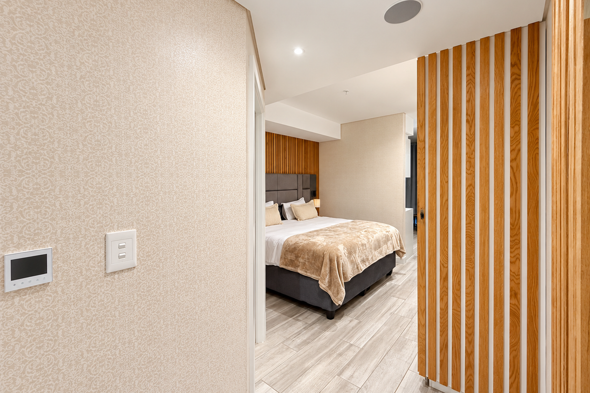 A modern bedroom featuring timber slat wall paneling, an upholstered headboard, recessed ceiling lighting, and wood-look tile flooring.