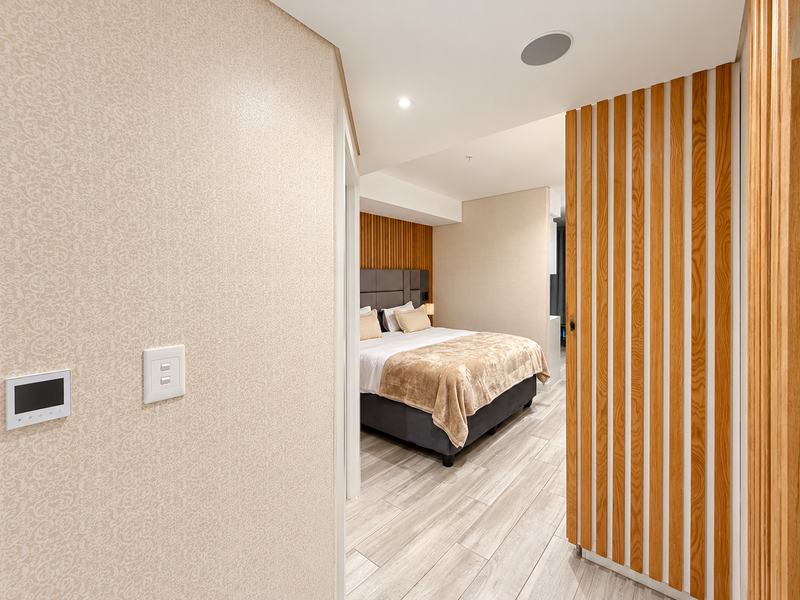 A modern bedroom featuring timber slat wall paneling, an upholstered headboard, recessed ceiling lighting, and wood-look tile flooring.