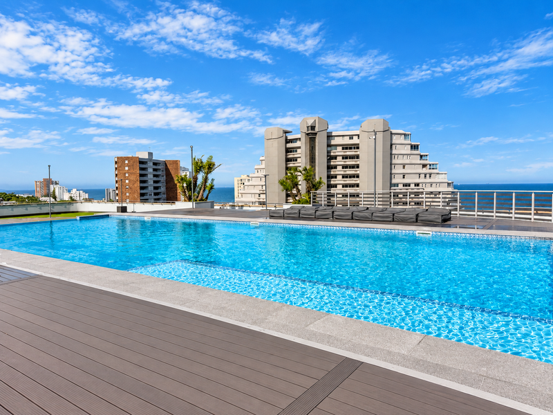 A well-maintained rooftop swimming pool featuring modern composite decking, stone coping, and panoramic ocean and city views.