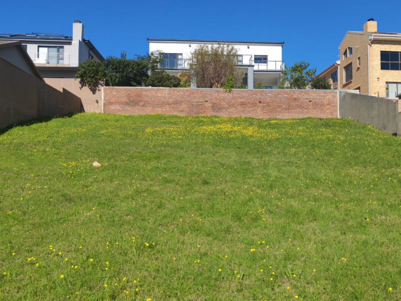 A sloped vacant residential plot featuring a brick retaining wall and established boundary walls, situated in a developed residential area.