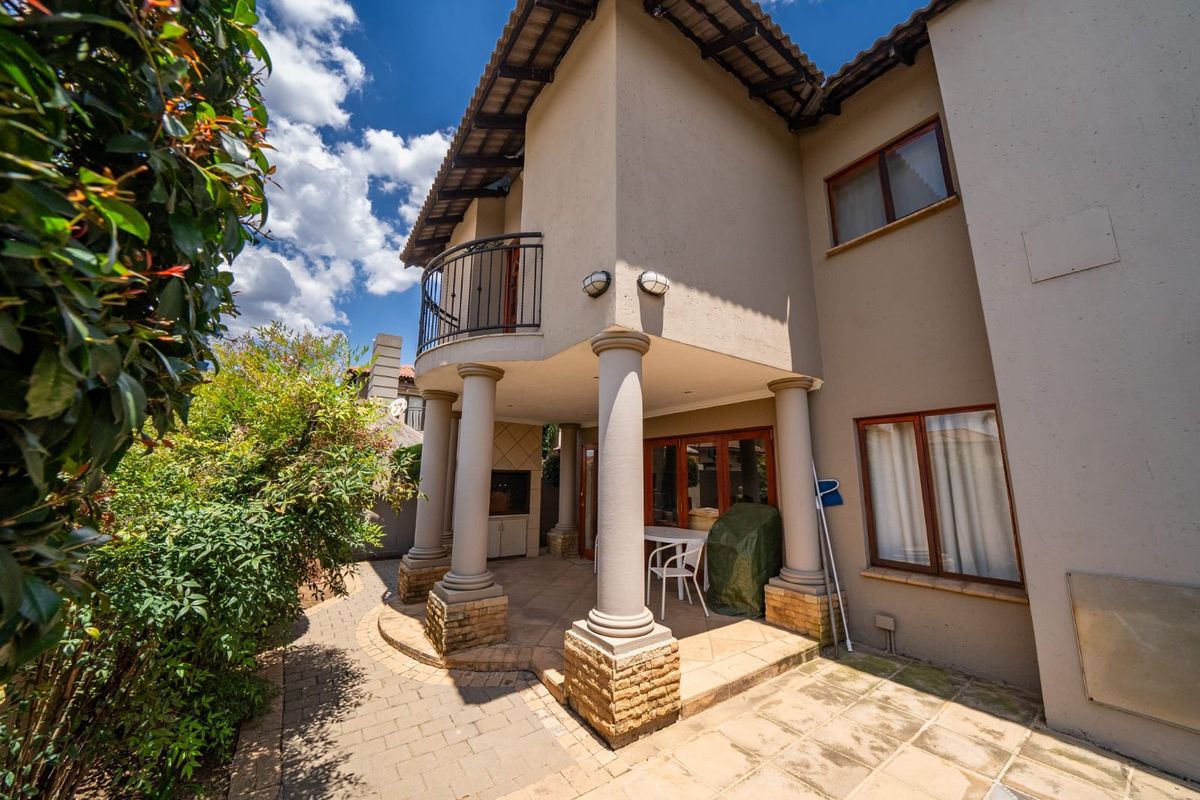 Rear view of the property featuring a covered patio with stone-clad columns, a built-in braai, and an upper-level balcony.
