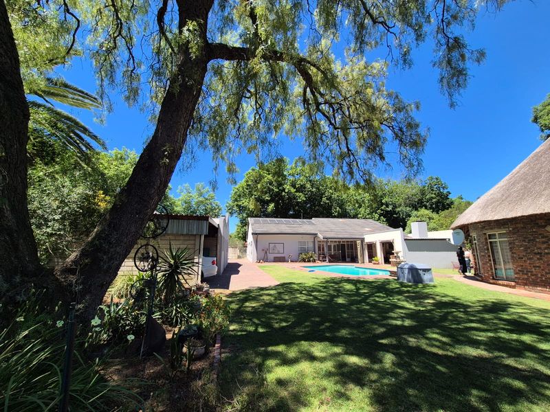 Spacious rear garden featuring a swimming pool, paved patio area, and a mix of brick and thatched roof structures with established trees.