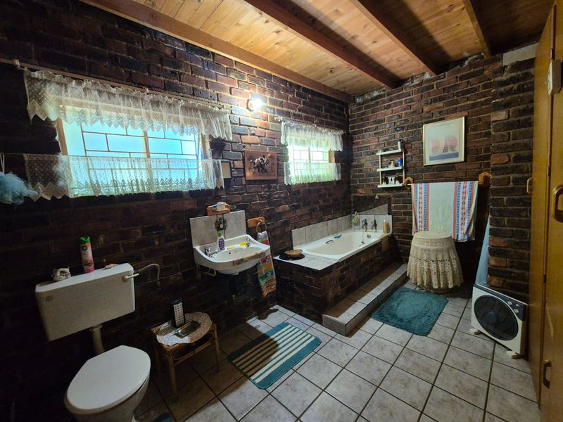 A rustic bathroom featuring exposed brick walls, timber beam ceiling, built-in bathtub, and wall-mounted basin with tiled flooring.