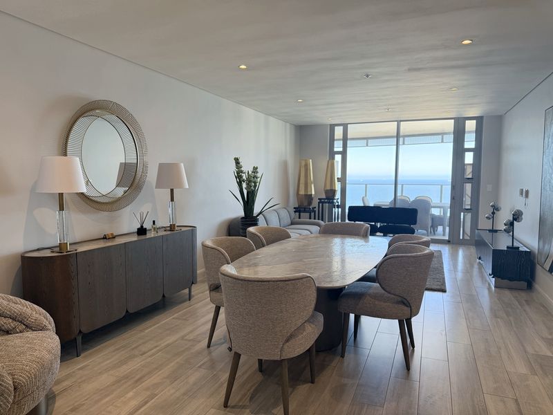 Spacious open-plan living and dining area featuring modern wood-look tile flooring, recessed lighting, and large sliding glass doors leading to a balcony with ocean views.