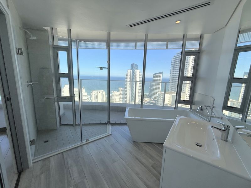 Modern bathroom featuring a freestanding bathtub, glass shower, and expansive floor-to-ceiling windows offering panoramic ocean and city views.
