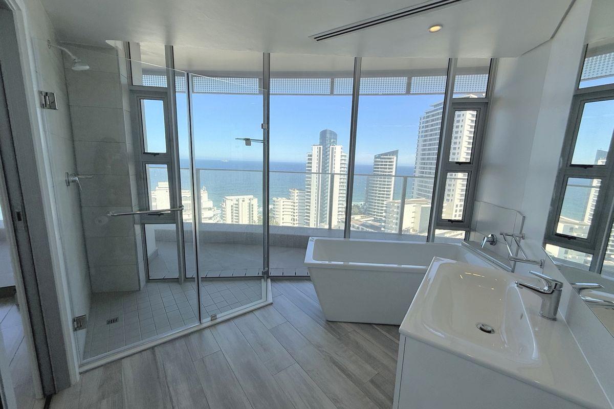 Modern bathroom featuring a freestanding bathtub, glass shower, and expansive floor-to-ceiling windows offering panoramic ocean and city views.