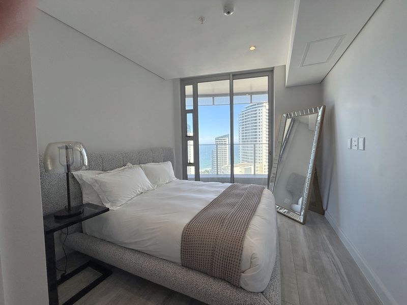 A modern, well-maintained bedroom featuring light-toned laminate flooring, recessed ceiling lighting, and a large sliding glass door providing access to a balcony with city views.