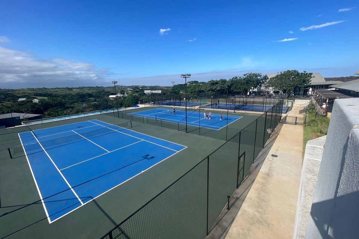 Elevated view of well-maintained blue tennis courts with perimeter fencing and floodlight infrastructure within a residential estate setting.