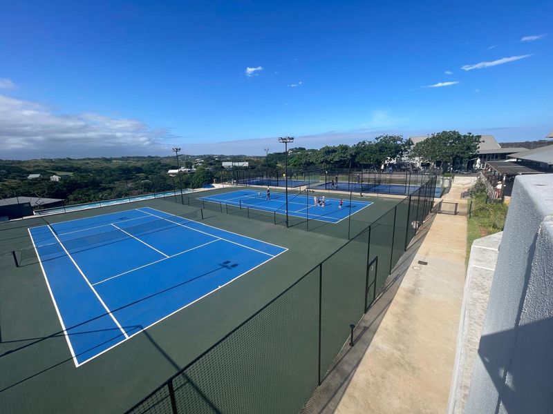 Elevated view of well-maintained blue tennis courts with perimeter fencing and floodlight infrastructure within a residential estate setting.