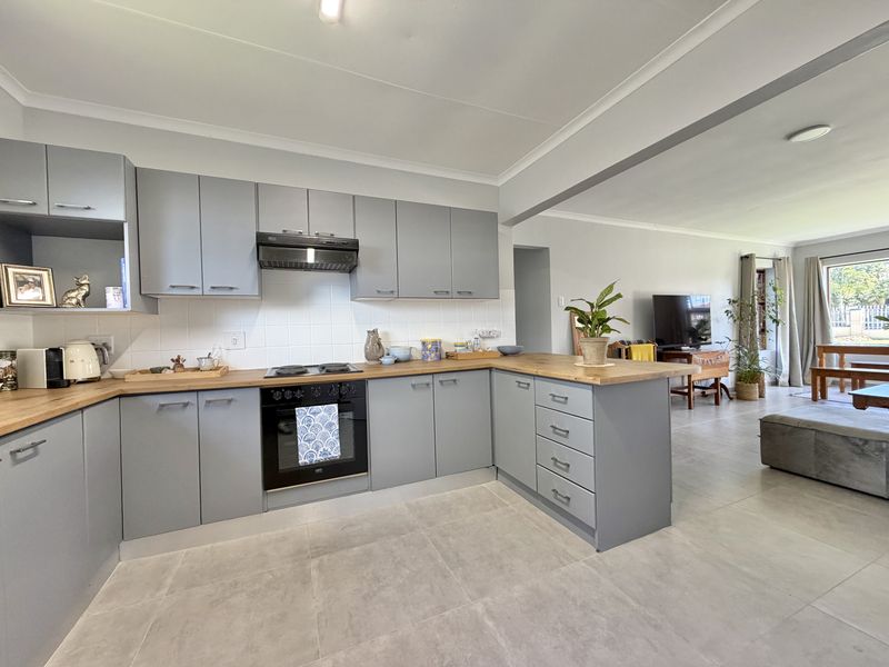 Modern open-plan kitchen featuring grey cabinetry, wooden countertops, built-in oven, and hob, seamlessly flowing into the adjacent living area with tiled flooring throughout.