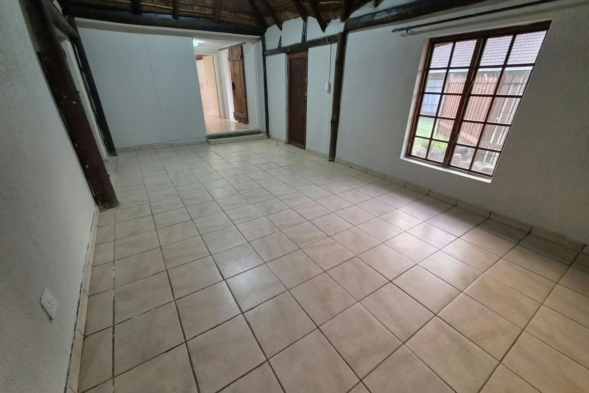 An empty living area featuring a rustic thatched roof with exposed timber beams, tiled flooring, and natural light through a timber-framed window.