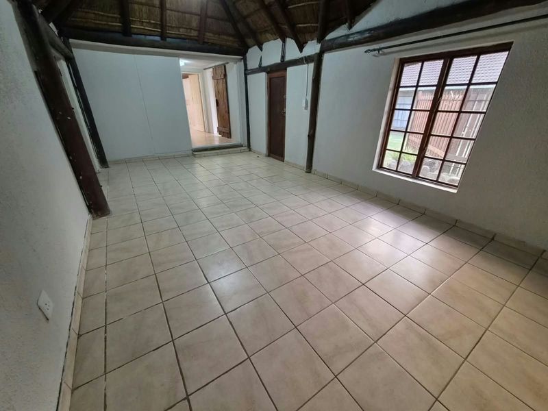 An empty living area featuring a rustic thatched roof with exposed timber beams, tiled flooring, and natural light through a timber-framed window.