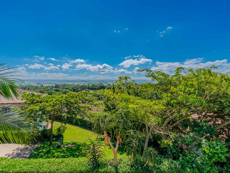 A scenic elevated view overlooking a lush green garden, mature trees, and a distant landscape under a clear blue sky.