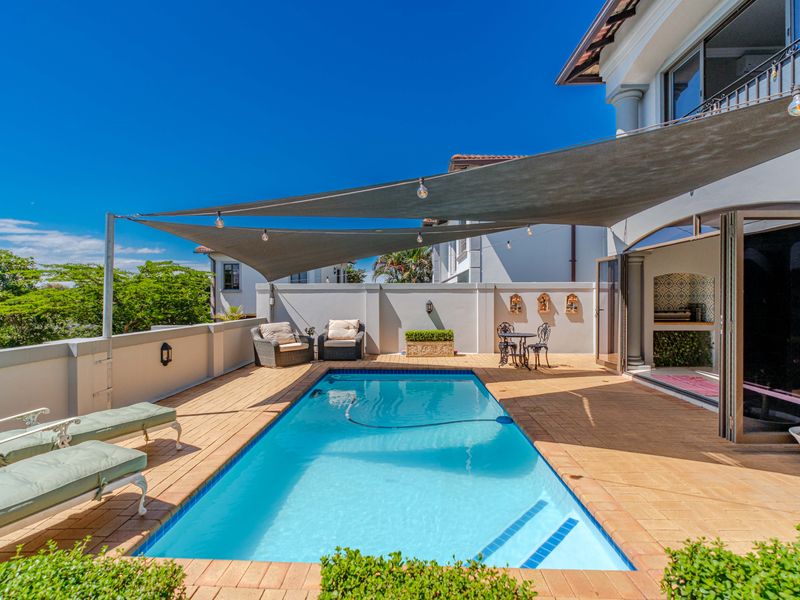 A well-maintained outdoor pool area featuring a paved deck, shade sail, and a built-in braai area with direct access to the home via bi-fold doors.