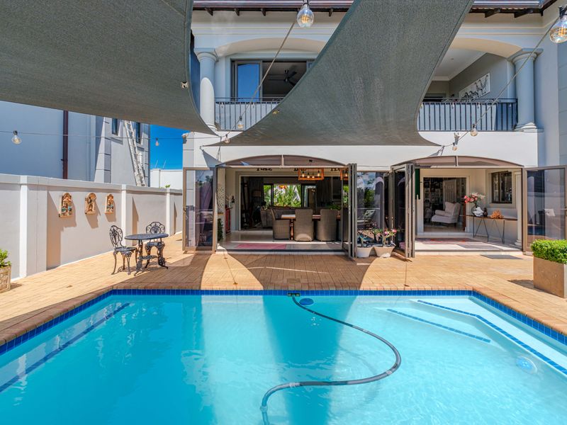 A well-maintained outdoor pool area featuring a paved deck, shade sail, and seamless indoor-outdoor flow through large bi-fold doors to the main residence.