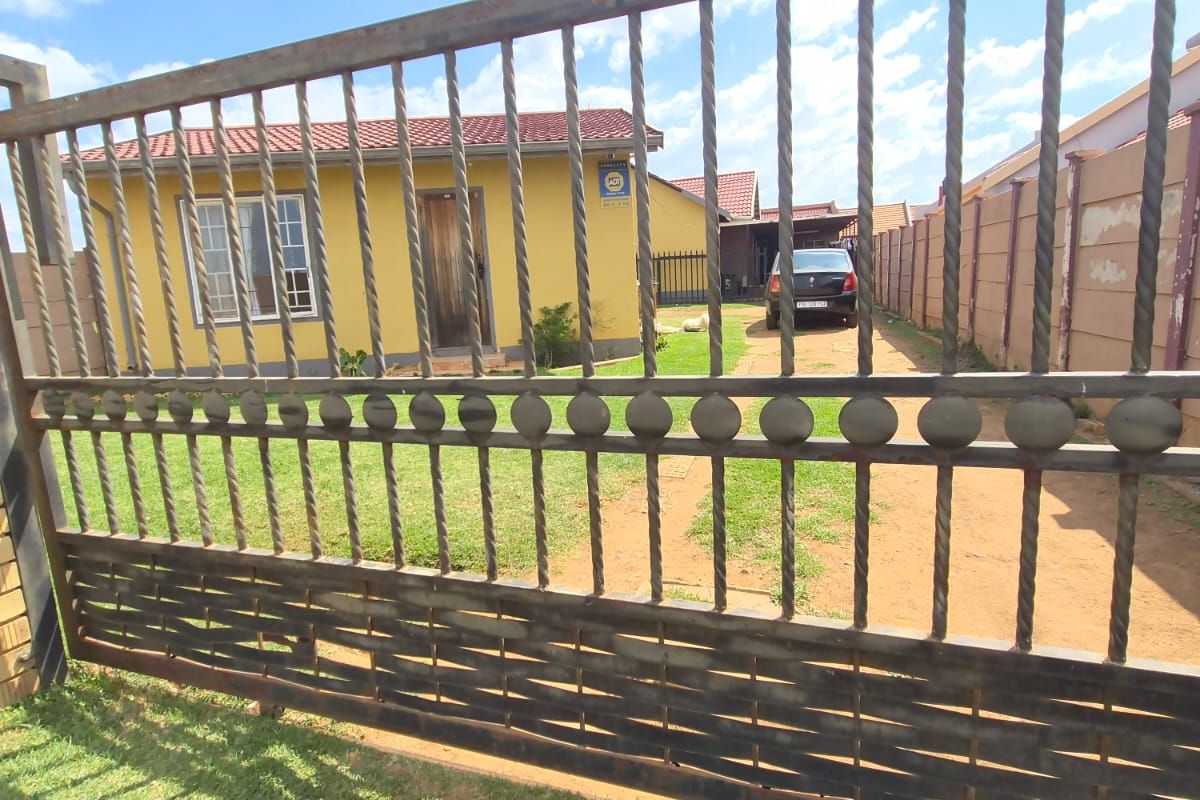 Front view of a residential property featuring a metal driveway gate, tiled roof, and a paved driveway leading to the house entrance.