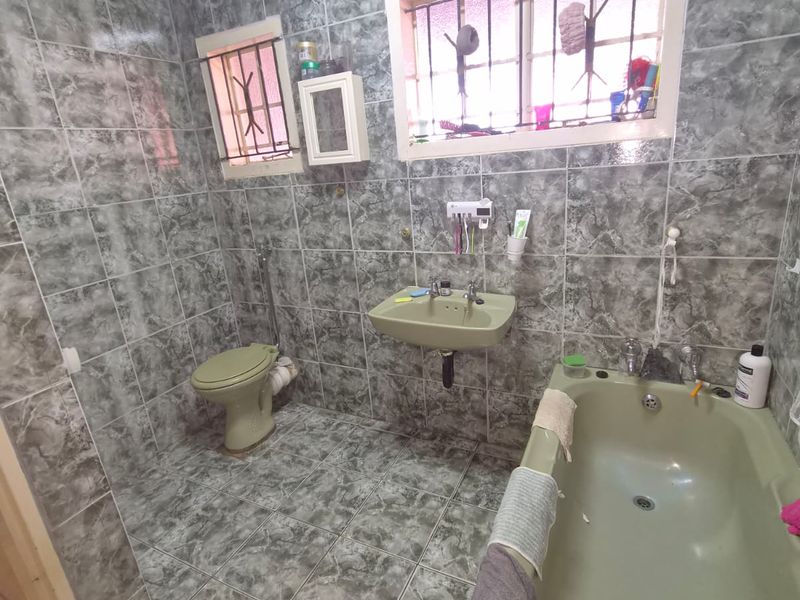 A dated bathroom featuring full-height grey marble-effect wall tiles, a green bathtub, matching basin, and toilet with security bars on the windows.