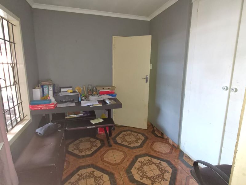 A compact office space featuring built-in cupboards, security bars on the window, and patterned tiled flooring, suitable for a home workspace.