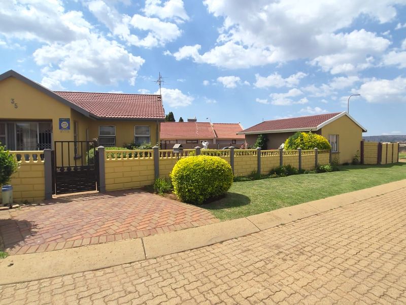 Front exterior view of a residential house featuring a paved driveway, brick boundary wall, and tiled roof under a clear sky.