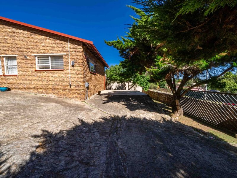 Exterior view of a face brick house featuring a concrete driveway, metal security fencing, and mature trees on a sunny day.