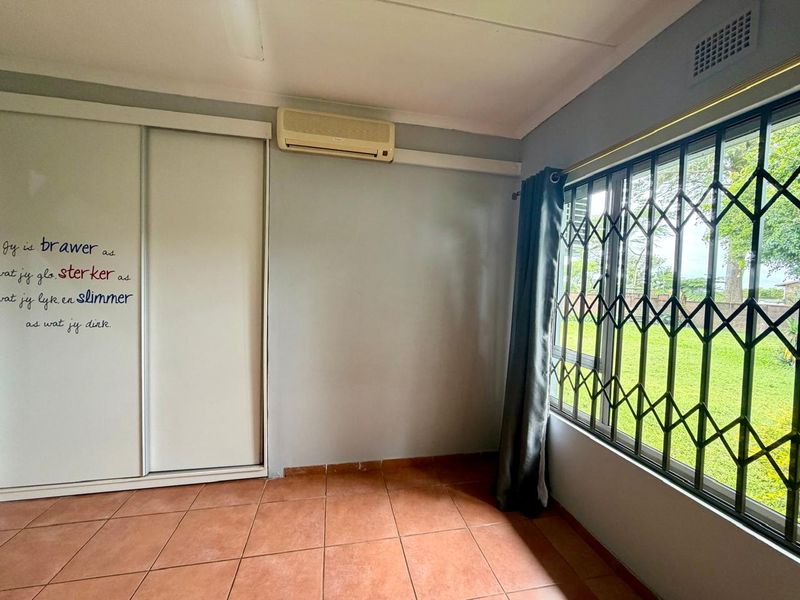 A bright, empty bedroom featuring tiled flooring, a built-in sliding wardrobe, and a wall-mounted air conditioning unit with security bars on the window.