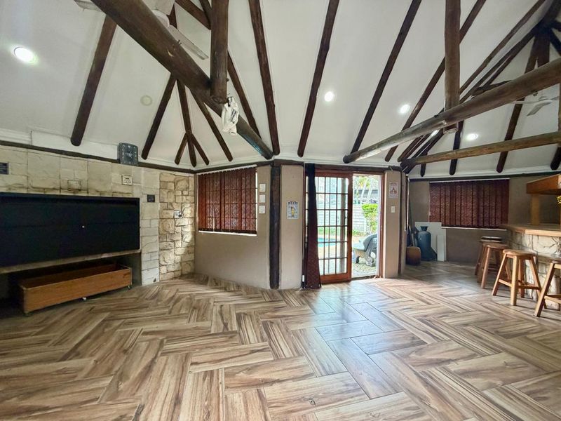 Spacious living area featuring exposed timber rafters, a stone-clad fireplace, and stylish herringbone floor tiles, with direct access to the outdoor pool area.