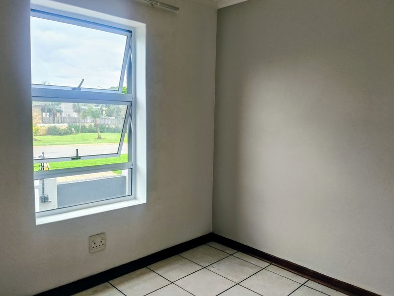 A bright, empty bedroom featuring tiled flooring, neutral walls, and an aluminium window providing natural light and a view of the exterior.