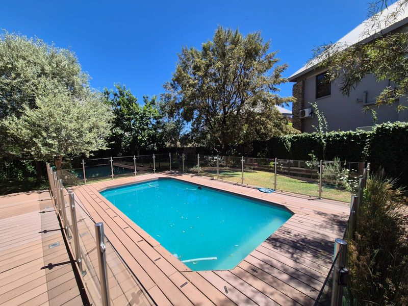 A well-maintained swimming pool area featuring a timber deck surround and modern glass balustrade, set within a landscaped garden.