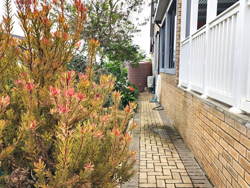 A side pathway featuring brick paving, a brick exterior wall, and a white balustrade, leading towards a rear garden area with a water tank.
