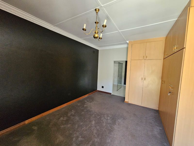 A spacious bedroom featuring built-in wooden cupboards, decorative ceiling cornicing, and a classic chandelier, with direct access to an en-suite bathroom.