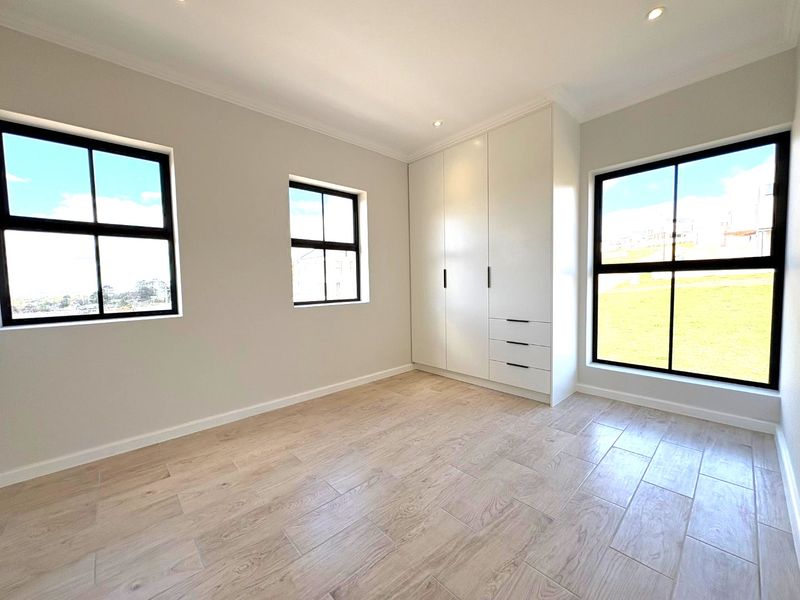 A bright, modern bedroom featuring neutral walls, wood-look tiled flooring, built-in cupboards, and large black-framed windows providing ample natural light.