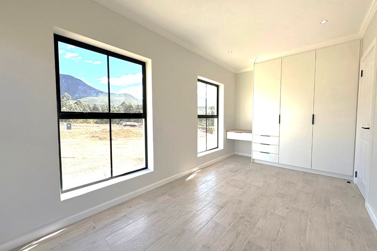 A bright, modern bedroom featuring wood-look tile flooring, built-in cupboards with an integrated desk, and large black-framed windows with scenic views.