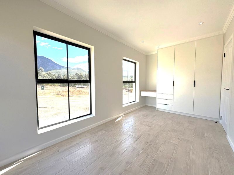 A bright, modern bedroom featuring wood-look tile flooring, built-in cupboards with an integrated desk, and large black-framed windows with scenic views.