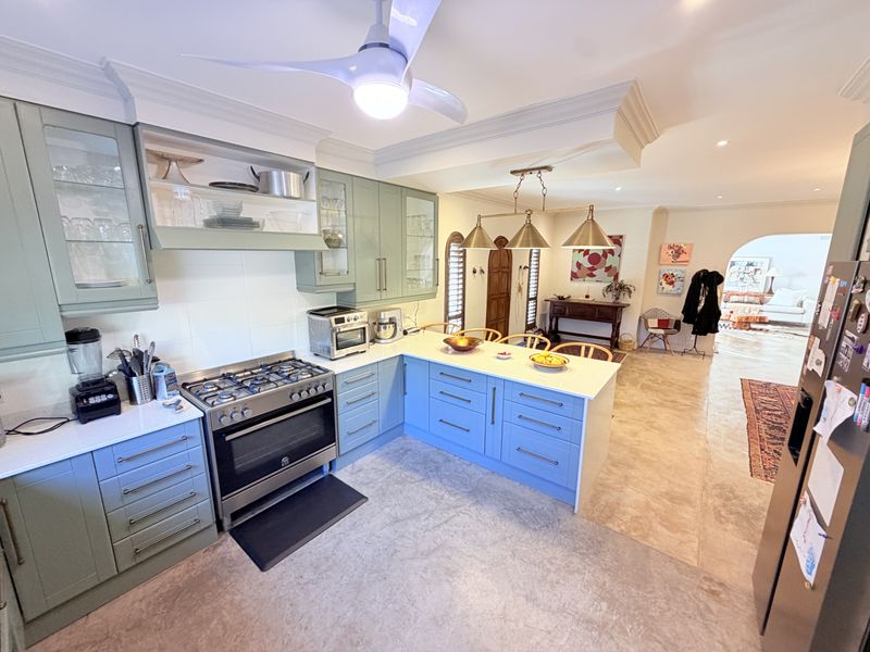 A well-appointed kitchen featuring sage green shaker cabinetry, stone countertops, a gas range, and integrated pendant lighting, flowing into an open-plan living area.